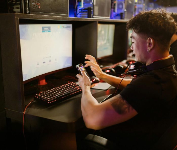 A young man is gaming using a smartphone and a PC setup with a mechanical keyboard indoors.