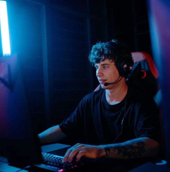 A young man intensely gaming with a headset at a computer desk, illuminated by blue light.